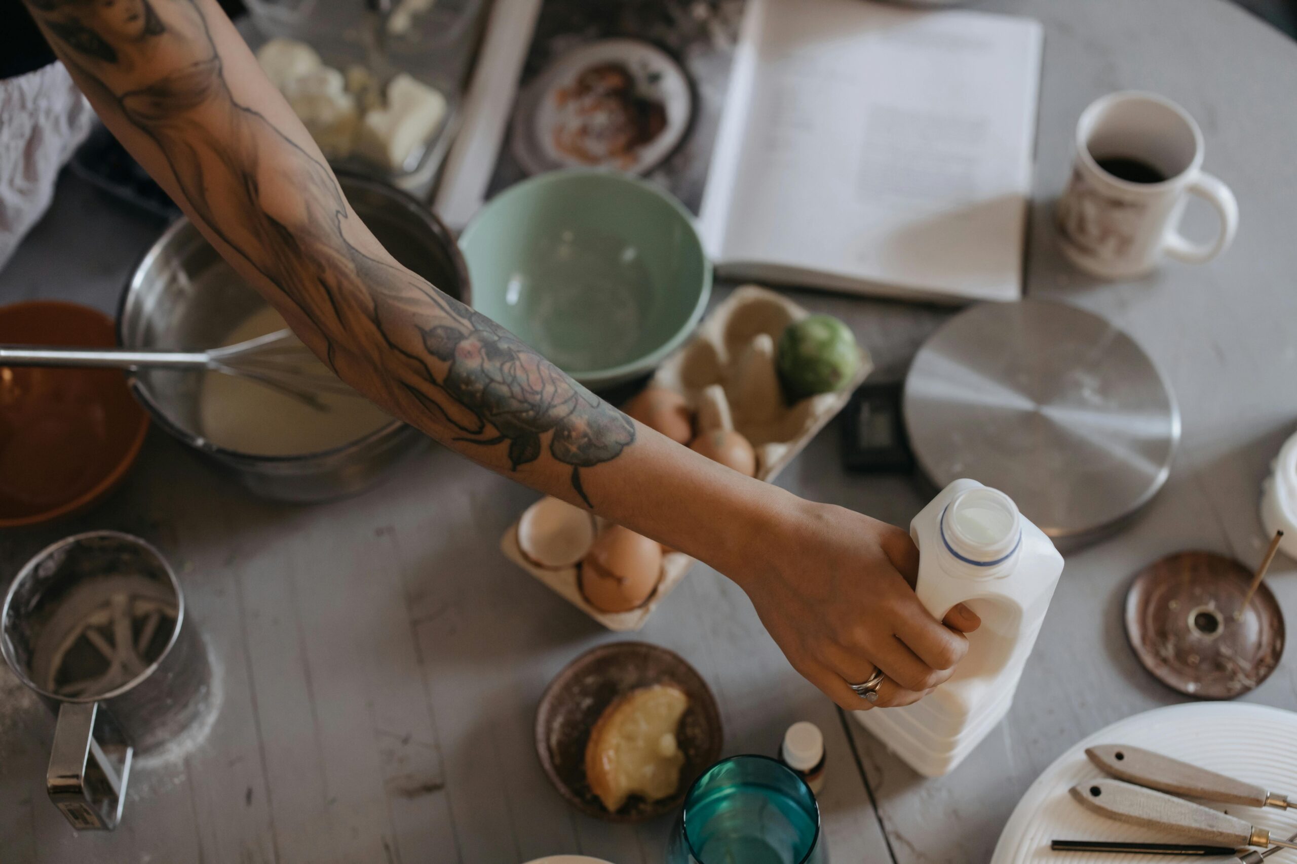 Tattooed person pouring milk in a rustic kitchen setting with variety of ingredients on table.